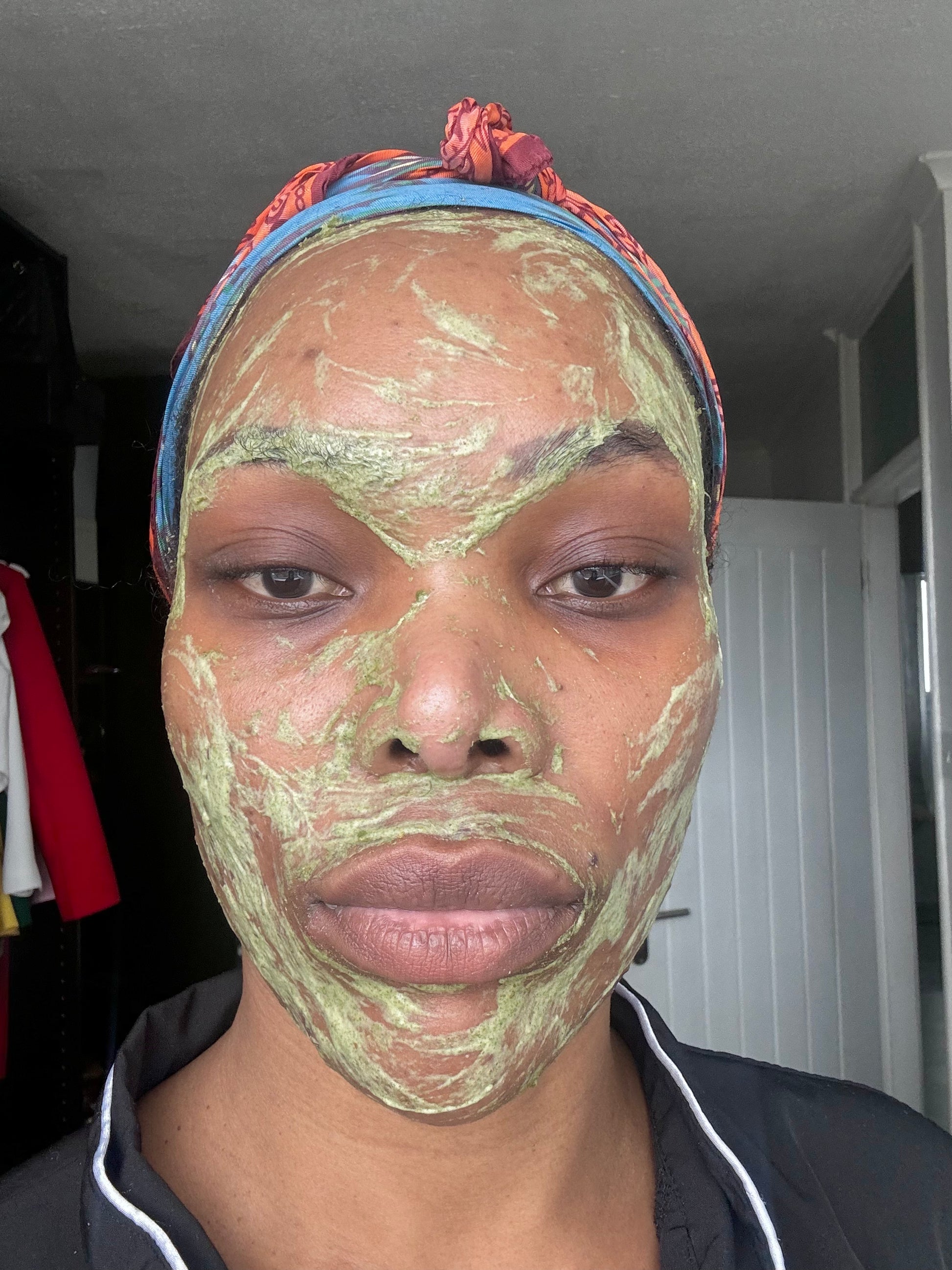A person with a green face mask applied evenly across their face, wearing a colourful headscarf and a black top. The face mask appears to be a natural skincare treatment, with a relaxed and calm expression.
