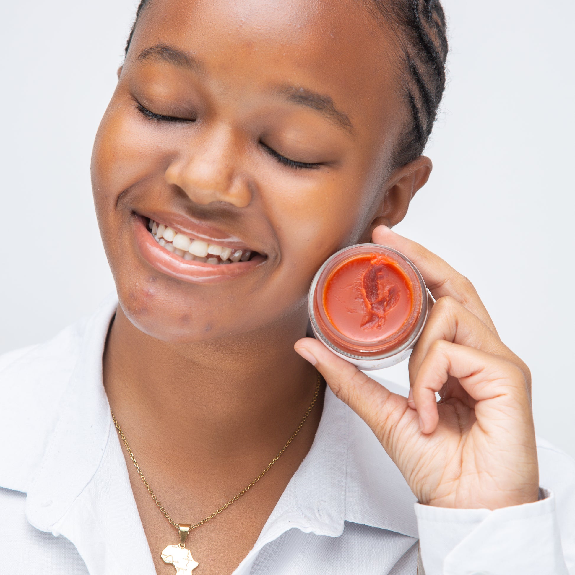 A young woman smiling with her eyes closed, holding a jar of vibrant orange skincare product. The product's smooth texture is visible in the jar, highlighting its rich, moisturizing formula. The woman looks happy and confident, emphasizsng the product's positive effect on her skin. She wears a gold Africa-shaped necklace, adding a personal and cultural touch to the image