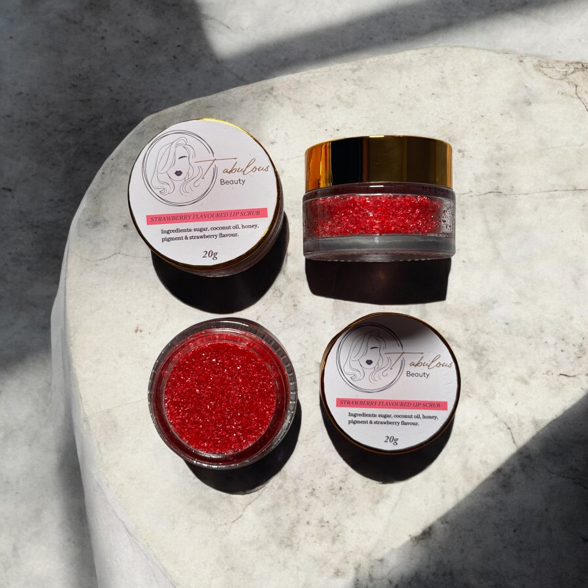 A top-down view of strawberry lip scrub jars arranged on a stone surface, with sunlight casting soft shadows. The rich red texture of the scrub sparkles inside the jars, highlighting the exfoliating and moisturising properties. The clean design of the Tabulous Beauty labels complements the natural beauty of the product, creating an inviting and luxurious feel